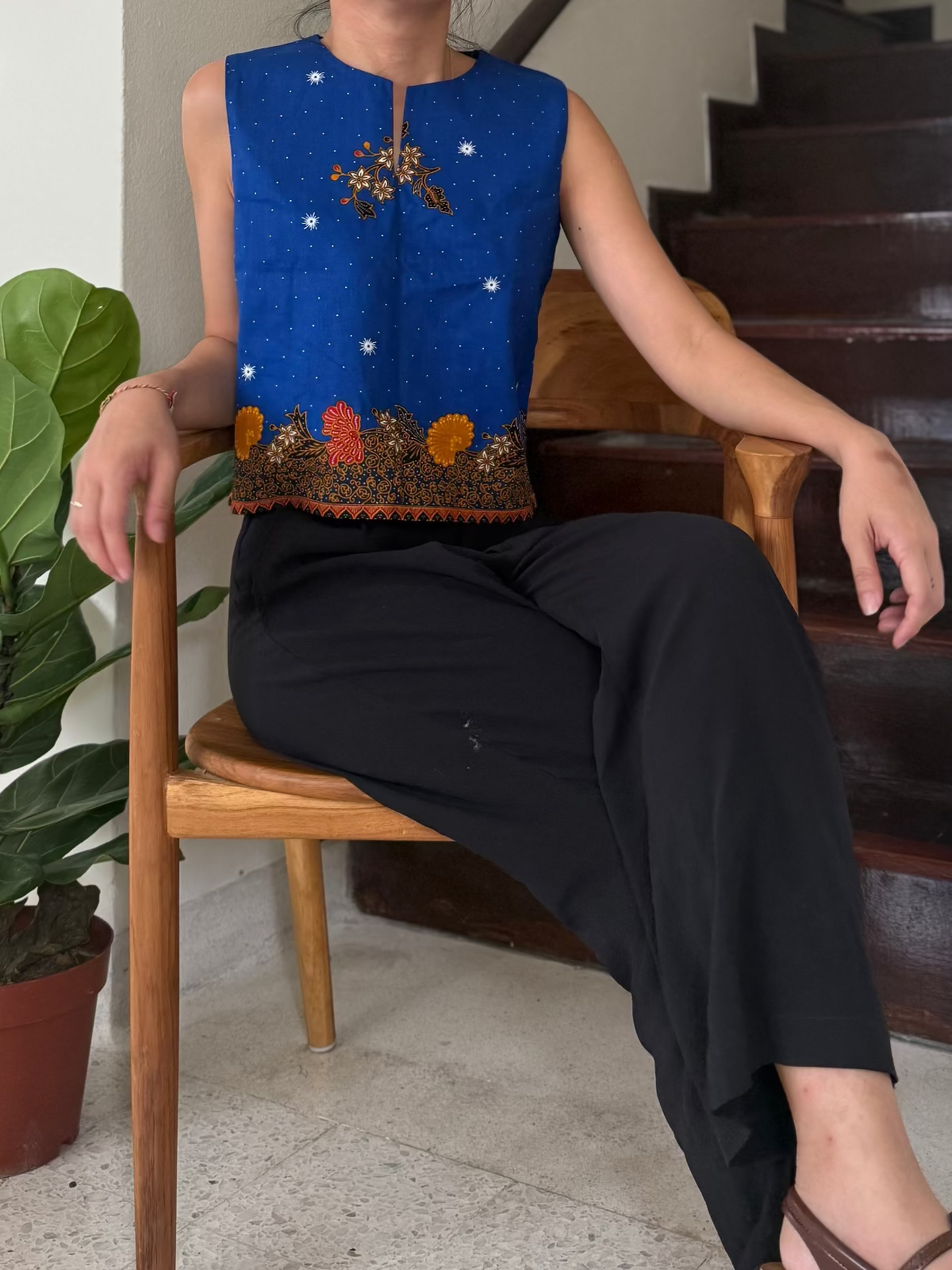 Person wearing a blue embroidered top and black pants sitting on a wooden chair.