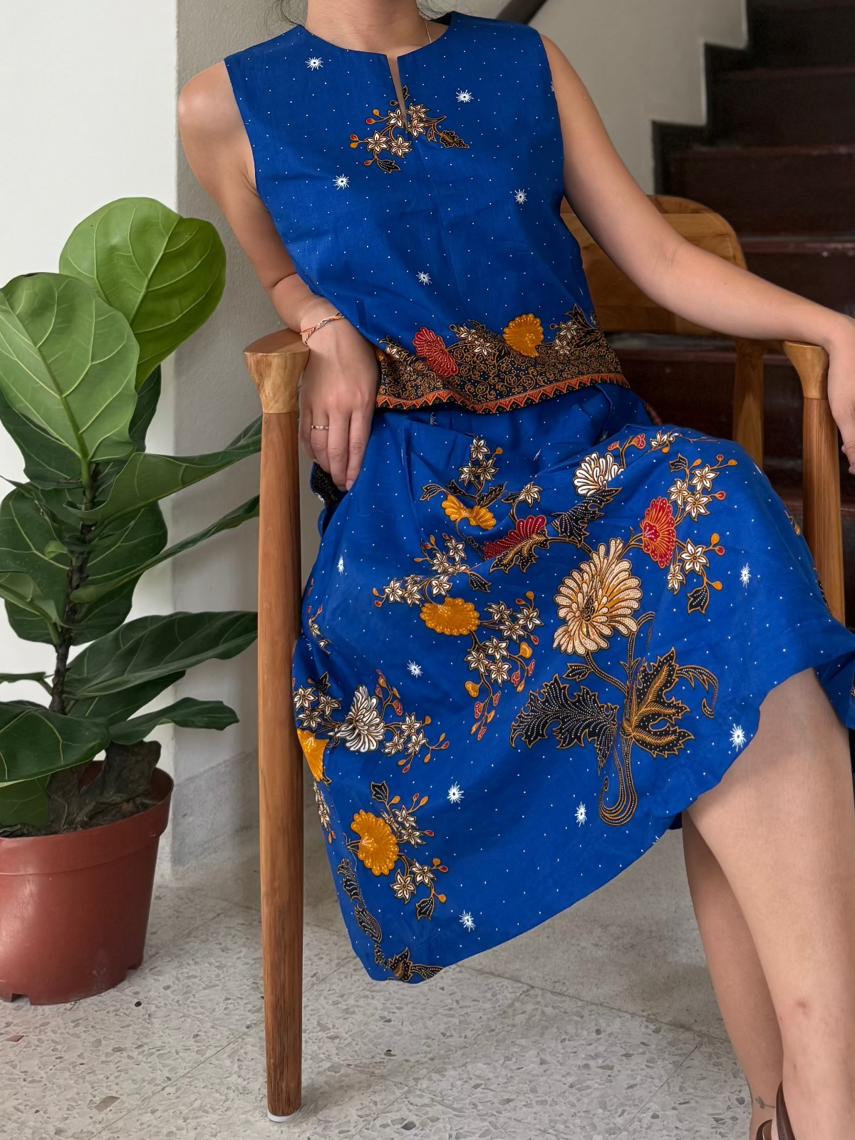Person wearing a blue embroidered dress sitting on a chair next to a potted plant.